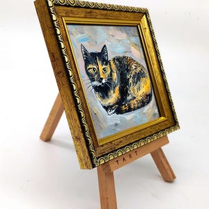 Pode incluir: Uma pequena pintura a &oacute;leo de um gato tartaruga numa moldura dourada ornamentada, exibida num cavalete de madeira. A pintura apresenta pinceladas vis&iacute;veis e a palavra TART est&aacute; impressa no cavalete de madeira clara.