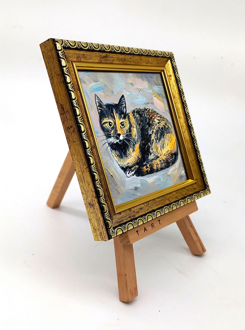 Pode incluir: Uma pequena pintura a &oacute;leo de um gato tartaruga numa moldura dourada ornamentada, exibida num cavalete de madeira. A pintura apresenta pinceladas vis&iacute;veis e a palavra TART est&aacute; impressa no cavalete de madeira clara.