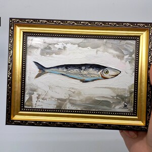 Puede incluir: Una pintura enmarcada de un pez plateado con marcas azules y negras sobre un fondo blanco con pinceladas grises y marrones. El marco es dorado con un borde exterior marr&oacute;n. La pintura est&aacute; firmada "JK" en la esquina inferior derecha.