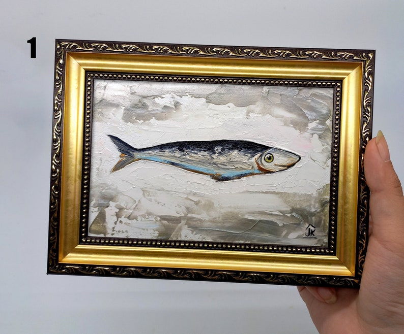 Puede incluir: Una pintura enmarcada de un pez plateado con marcas azules y negras sobre un fondo blanco con pinceladas grises y marrones. El marco es dorado con un borde exterior marr&oacute;n. La pintura est&aacute; firmada "JK" en la esquina inferior derecha.