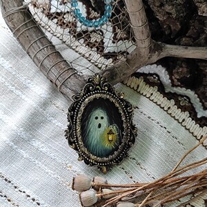 May include: An oval antique-style frame featuring a painting of a ghost holding a lantern. The ghost is white with a surprised expression. The frame is dark with gold accents. A blue beaded bracelet and dried seed pods are also visible.