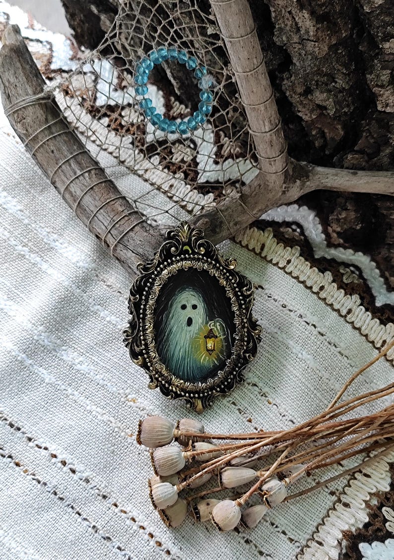 May include: An oval antique-style frame featuring a painting of a ghost holding a lantern. The ghost is white with a surprised expression. The frame is dark with gold accents. A blue beaded bracelet and dried seed pods are also visible.