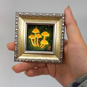 May include: A framed miniature painting of a cluster of glowing yellow mushrooms on a dark green background with white speckles. The painting is signed "JR" in the bottom right corner.
