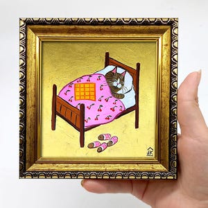 Puede incluir: Una pintura enmarcada de un gato durmiendo en una cama rosa con estampado de cerezas. El gato es marr&oacute;n y blanco y tiene los ojos cerrados. La cama tiene una manta a cuadros amarilla encima. Hay dos pares de zapatillas rosas y blancas en el suelo al lado de la cama. La pintura est&aacute; sobre un fondo dorado y tiene un marco dorado.