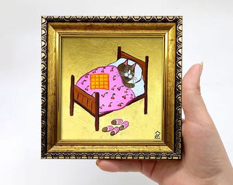 May include: A framed painting of a cat sleeping in a pink bed with a cherry pattern. The cat is brown and white and has its eyes closed. The bed has a yellow checkered blanket on top of it. There are two pairs of pink and white slippers on the floor next to the bed. The painting is on a gold background and has a gold frame.