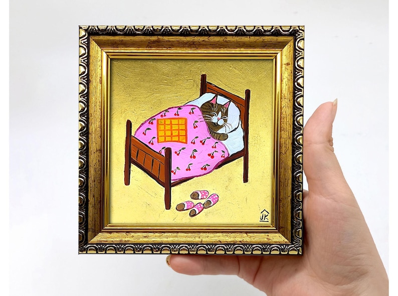 Puede incluir: Una pintura enmarcada de un gato durmiendo en una cama rosa con estampado de cerezas. El gato es marr&oacute;n y blanco y tiene los ojos cerrados. La cama tiene una manta a cuadros amarilla encima. Hay dos pares de zapatillas rosas y blancas en el suelo al lado de la cama. La pintura est&aacute; sobre un fondo dorado y tiene un marco dorado.