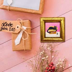 May include: A black cat sits in a pink and white box with the word "PARENTS" written on it. The cat is looking to the right. The box is on a pink background. The image is framed in gold.