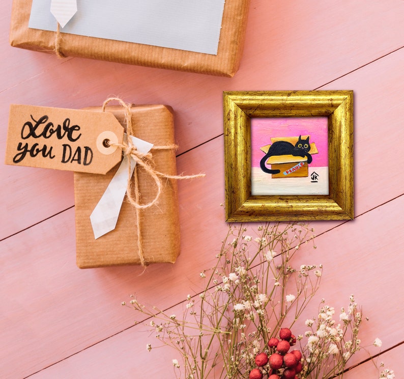 May include: A black cat sits in a pink and white box with the word "PARENTS" written on it. The cat is looking to the right. The box is on a pink background. The image is framed in gold.