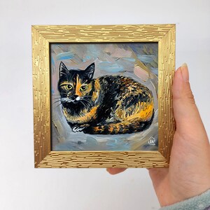 Pode incluir: Uma pequena pintura a &oacute;leo quadrada de um gato tartaruga com pelagem preta, laranja e branca, olhos amarelos e bigodes longos. A obra est&aacute; em uma moldura dourada texturizada com um padr&atilde;o de tra&ccedil;os horizontais repetidos e a assinatura JK.