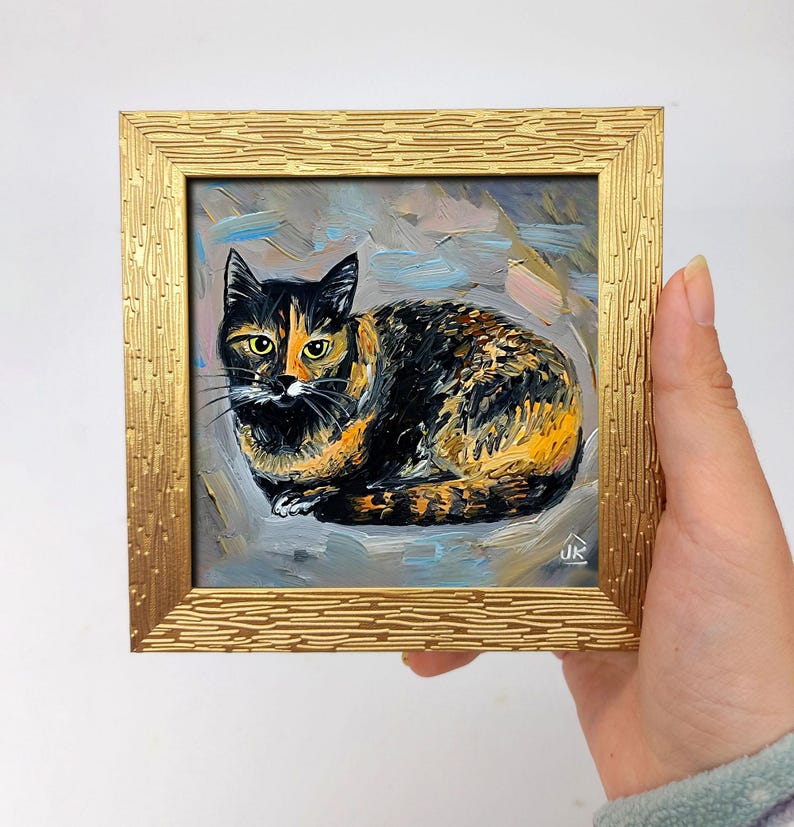 Pode incluir: Uma pequena pintura a &oacute;leo quadrada de um gato tartaruga com pelagem preta, laranja e branca, olhos amarelos e bigodes longos. A obra est&aacute; em uma moldura dourada texturizada com um padr&atilde;o de tra&ccedil;os horizontais repetidos e a assinatura JK.
