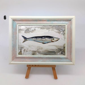 Puede incluir: Una pintura enmarcada de un pez plateado con una franja azul sobre un fondo blanco con pinceladas grises y marrones. El marco es blanco con un lavado azul claro y rosa. La pintura est&aacute; en un caballete con la palabra "TART" escrita en &eacute;l.