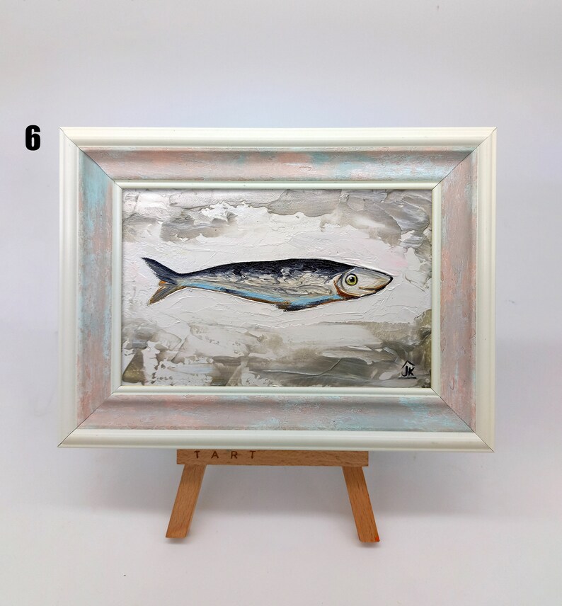 Puede incluir: Una pintura enmarcada de un pez plateado con una franja azul sobre un fondo blanco con pinceladas grises y marrones. El marco es blanco con un lavado azul claro y rosa. La pintura est&aacute; en un caballete con la palabra "TART" escrita en &eacute;l.