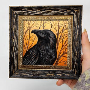 Pode incluir: Pintura emoldurada de um corvo preto com detalhes de penas, em um fundo laranja e marrom de &aacute;rvores nuas. A moldura &eacute; preta com detalhes dourados.