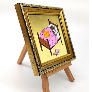 Puede incluir: Una pintura enmarcada de un gato durmiendo en una cama con una manta rosa con estampado de cerezas. La cama es de madera marr&oacute;n y el gato es gris y blanco. La pintura est&aacute; en un peque&ntilde;o caballete de madera con la palabra "TART" escrita en la parte inferior.