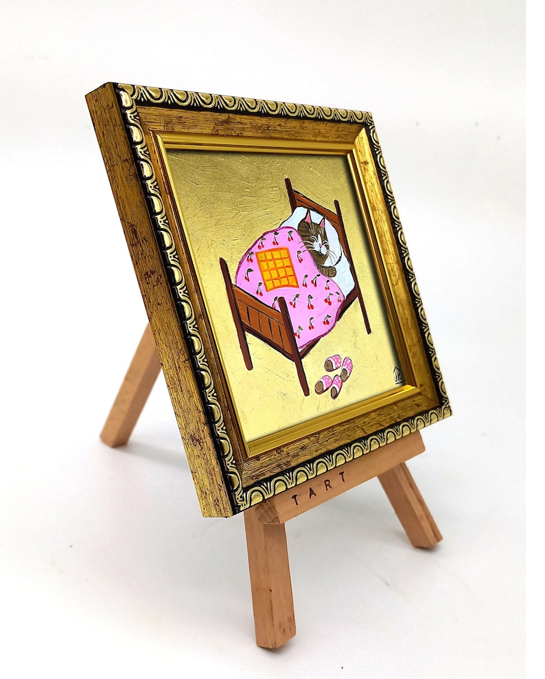 Puede incluir: Una pintura enmarcada de un gato durmiendo en una cama con una manta rosa con estampado de cerezas. La cama es de madera marr&oacute;n y el gato es gris y blanco. La pintura est&aacute; en un peque&ntilde;o caballete de madera con la palabra "TART" escrita en la parte inferior.