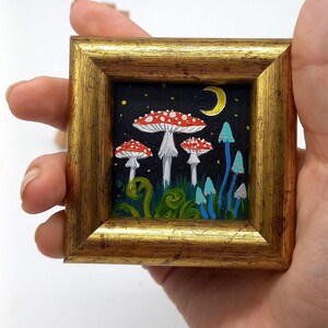 May include: A framed painting depicting a whimsical forest scene with red and white mushrooms, blue mushrooms, and a crescent moon in a starry night sky.