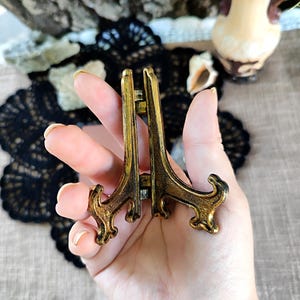 May include: A small, antique-style brass display stand with an ornate design. The stand is held in a hand, showcasing its intricate details and aged finish. The stand is designed to hold small decorative items.