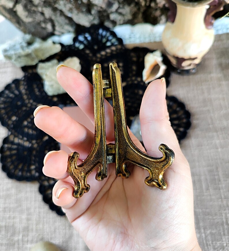 May include: A small, antique-style brass display stand with an ornate design. The stand is held in a hand, showcasing its intricate details and aged finish. The stand is designed to hold small decorative items.