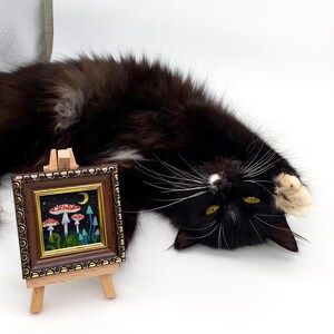 May include: A small framed painting on an easel depicts a whimsical scene of red and white mushrooms growing in a forest with a crescent moon in the night sky.