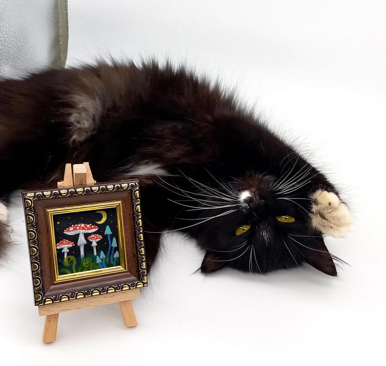 May include: A small framed painting on an easel depicts a whimsical scene of red and white mushrooms growing in a forest with a crescent moon in the night sky.