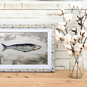 Puede incluir: Una pintura enmarcada de un pez plateado sobre un fondo blanco texturizado. El marco es blanco con detalles dorados. La pintura est&aacute; sobre una superficie de madera con un jarr&oacute;n de flores de algod&oacute;n blanco en el fondo.