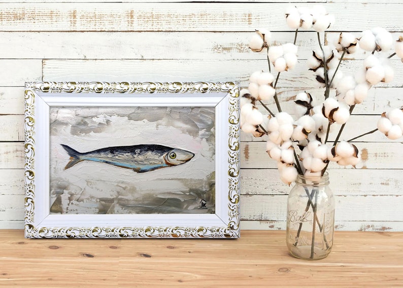Puede incluir: Una pintura enmarcada de un pez plateado sobre un fondo blanco texturizado. El marco es blanco con detalles dorados. La pintura est&aacute; sobre una superficie de madera con un jarr&oacute;n de flores de algod&oacute;n blanco en el fondo.