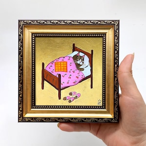 Puede incluir: Una pintura enmarcada de un gato durmiendo en una cama con una manta rosa con estampado de cerezas. El gato es marr&oacute;n y blanco y tiene los ojos cerrados. La cama es de madera y tiene una cabecera dorada. Hay dos pares de calcetines en el suelo al lado de la cama.