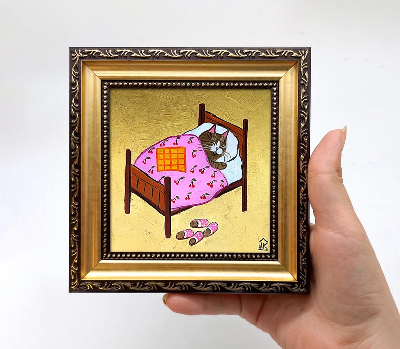 Puede incluir: Una pintura enmarcada de un gato durmiendo en una cama con una manta rosa con estampado de cerezas. El gato es marr&oacute;n y blanco y tiene los ojos cerrados. La cama es de madera y tiene una cabecera dorada. Hay dos pares de calcetines en el suelo al lado de la cama.