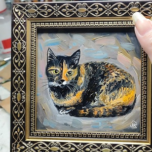 Pode incluir: Uma pintura a &oacute;leo de um gato tartaruga com pelagem preta e laranja e olhos amarelos, sobre um fundo cinza texturizado. A obra de arte est&aacute; emoldurada em uma moldura ornamentada marrom escura e dourada com padr&otilde;es de filigrana complexos.