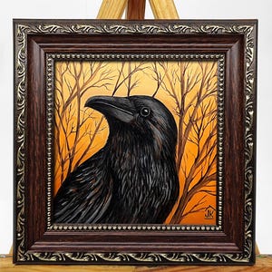 Pode incluir: Pintura emoldurada de um corvo preto com penas detalhadas, contra um fundo de &aacute;rvores nuas e c&eacute;u laranja. A obra de arte est&aacute; encerrada em uma moldura marrom escura e ornamentada com detalhes dourados, exibida em um cavalete de madeira.