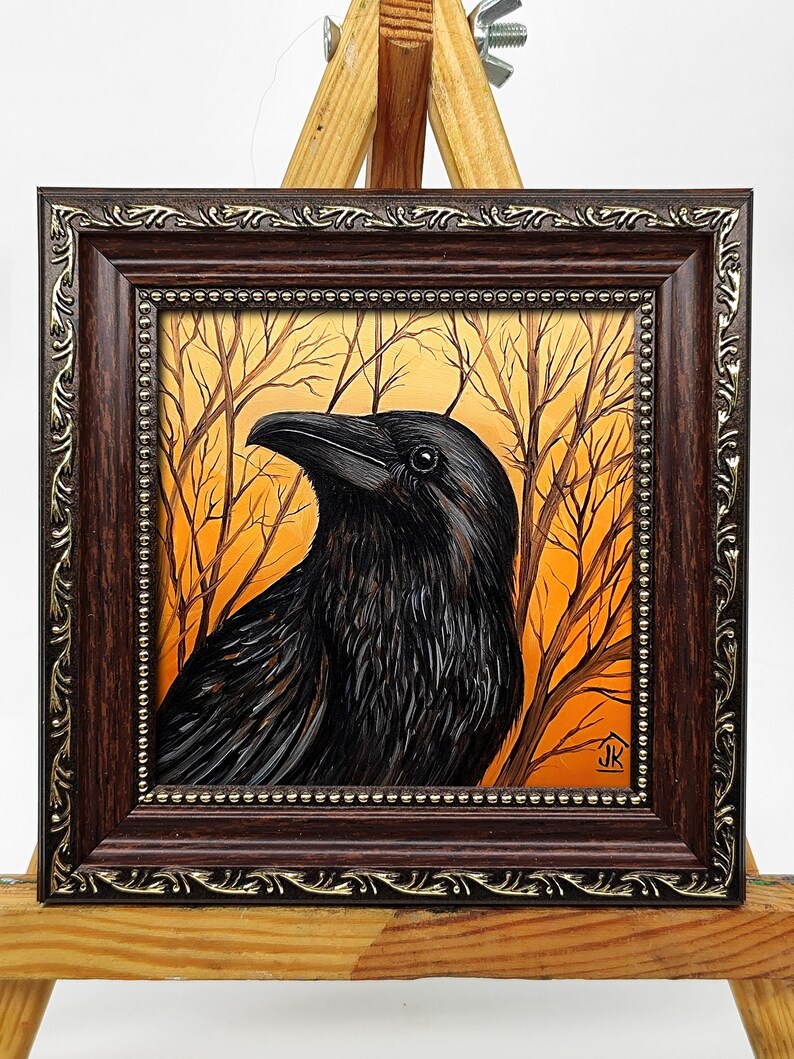 Pode incluir: Pintura emoldurada de um corvo preto com penas detalhadas, contra um fundo de &aacute;rvores nuas e c&eacute;u laranja. A obra de arte est&aacute; encerrada em uma moldura marrom escura e ornamentada com detalhes dourados, exibida em um cavalete de madeira.