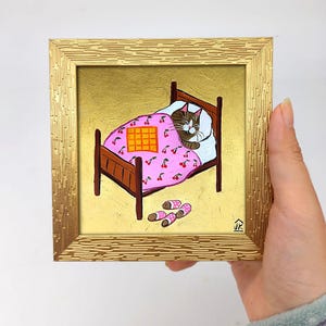Puede incluir: Una pintura enmarcada de un gato durmiendo en una cama con una manta rosa con estampado de cerezas. El gato es marr&oacute;n y blanco con una manta a cuadros amarilla y naranja encima. Hay zapatillas rosas y blancas en el suelo junto a la cama.