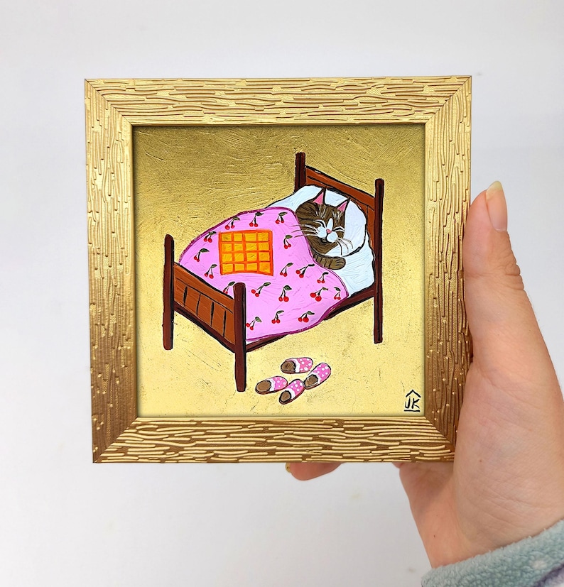 Puede incluir: Una pintura enmarcada de un gato durmiendo en una cama con una manta rosa con estampado de cerezas. El gato es marr&oacute;n y blanco con una manta a cuadros amarilla y naranja encima. Hay zapatillas rosas y blancas en el suelo junto a la cama.