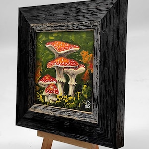 May include: A framed painting of red and white mushrooms in a forest setting. The painting is on a small wooden easel.