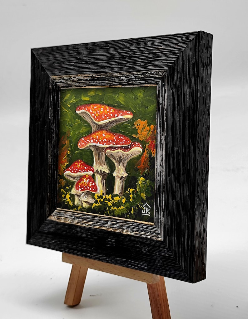 May include: A framed painting of red and white mushrooms in a forest setting. The painting is on a small wooden easel.