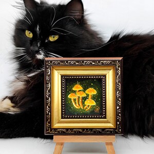 May include: A small framed painting of a cluster of yellow mushrooms on a green background with white speckles. The painting is on a wooden easel and a black cat is looking at it.