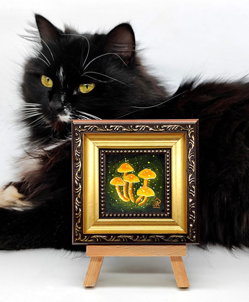 May include: A small framed painting of a cluster of yellow mushrooms on a green background with white speckles. The painting is on a wooden easel and a black cat is looking at it.