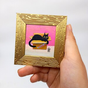 May include: A black cat with yellow eyes is lying in a cardboard box with a "FRAGILE" label. The box is on a pink surface and the cat is looking at the viewer. The painting is framed in a gold frame.