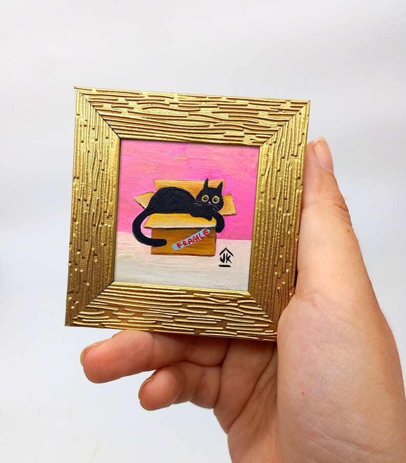 May include: A black cat with yellow eyes is lying in a cardboard box with a "FRAGILE" label. The box is on a pink surface and the cat is looking at the viewer. The painting is framed in a gold frame.