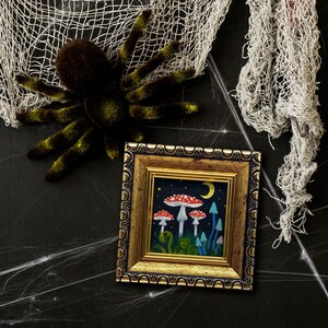 May include: A framed painting depicting a whimsical forest scene with mushrooms, a crescent moon, and stars. The mushrooms are red and white with white spots, and the background is a dark blue.