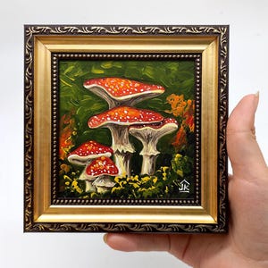May include: A framed painting of red and white mushrooms in a forest setting. The mushrooms are painted in a realistic style with a textured brushstroke. The painting is framed in a gold and brown frame.