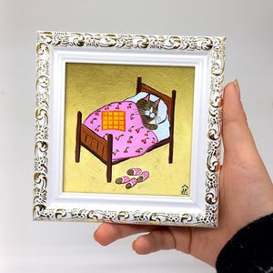 Puede incluir: Una pintura enmarcada de un gato durmiendo en una cama rosa y blanca con una manta a cuadros. La cama est&aacute; sobre un fondo dorado y hay zapatillas en el suelo.