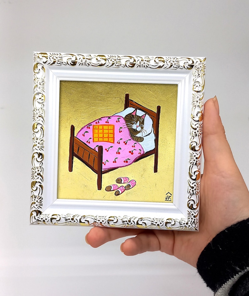 Puede incluir: Una pintura enmarcada de un gato durmiendo en una cama rosa y blanca con una manta a cuadros. La cama est&aacute; sobre un fondo dorado y hay zapatillas en el suelo.