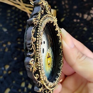 May include: An ornate, dark-framed pendant featuring a miniature painting of a ghostly figure holding a lantern. The frame has gold-coloured filigree detailing. The painting shows a white ghost with a yellow and orange lantern against a dark background.