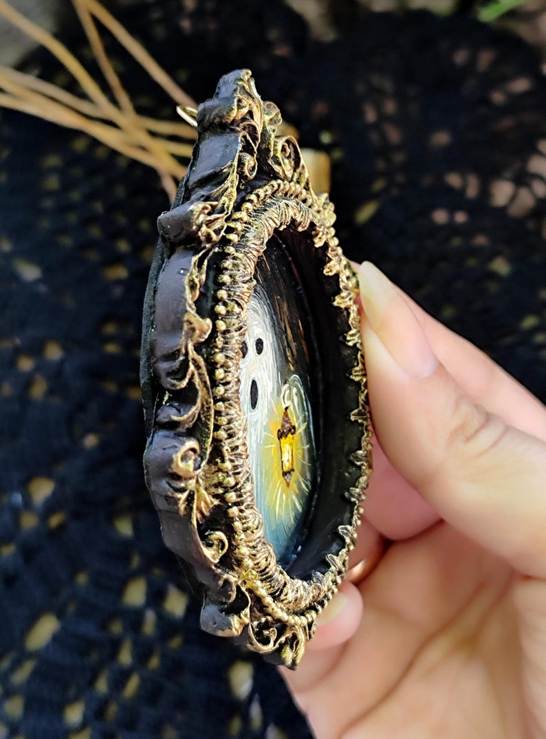 May include: An ornate, dark-framed pendant featuring a miniature painting of a ghostly figure holding a lantern. The frame has gold-coloured filigree detailing. The painting shows a white ghost with a yellow and orange lantern against a dark background.