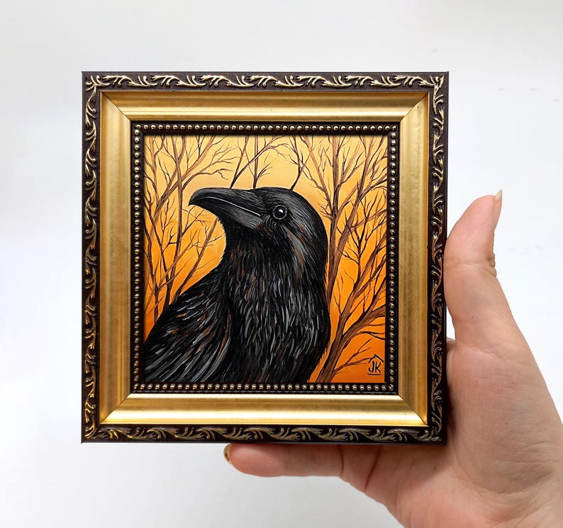 May include: A framed painting of a black raven with a detailed, realistic style. The raven is perched in a forest setting with a golden sunset background. The frame is ornate with a gold finish and a dark brown border.