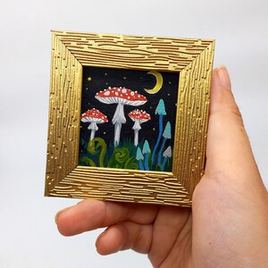 May include: A framed painting depicting a whimsical forest scene at night. The painting features three red and white mushrooms with white stems and a crescent moon in the background. The mushrooms are surrounded by green foliage and blue glowing mushrooms.