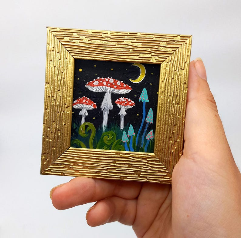 May include: A framed painting depicting a whimsical forest scene at night. The painting features three red and white mushrooms with white stems and a crescent moon in the background. The mushrooms are surrounded by green foliage and blue glowing mushrooms.