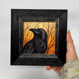 Pode incluir: Pintura emoldurada de um corvo preto com penas detalhadas, contra um fundo de &aacute;rvores nuas e c&eacute;u laranja. A obra de arte est&aacute; contida em uma moldura de madeira preta texturizada, criando um contraste marcante.
