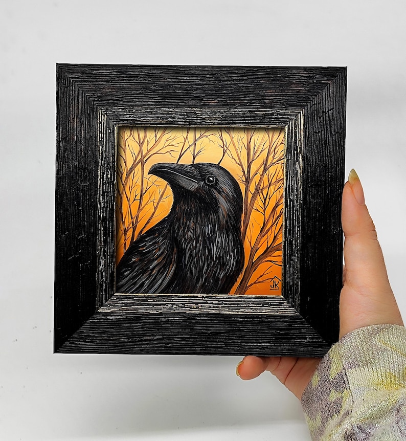 Pode incluir: Pintura emoldurada de um corvo preto com penas detalhadas, contra um fundo de &aacute;rvores nuas e c&eacute;u laranja. A obra de arte est&aacute; contida em uma moldura de madeira preta texturizada, criando um contraste marcante.
